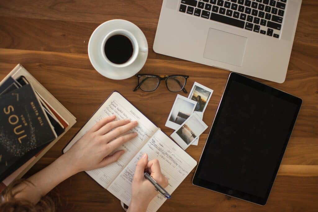 A notebook being written in on a desk with an iPad, some small photos, a cup of coffee, glasses, some books and a laptop there as well, the best calendar app would help.