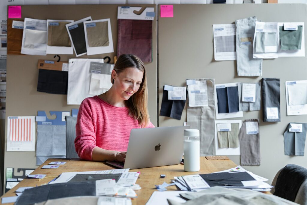 A person sits at a desk with many clothing samples on a table and behind them as they use their laptop giving a presentation using the best work from home apps.