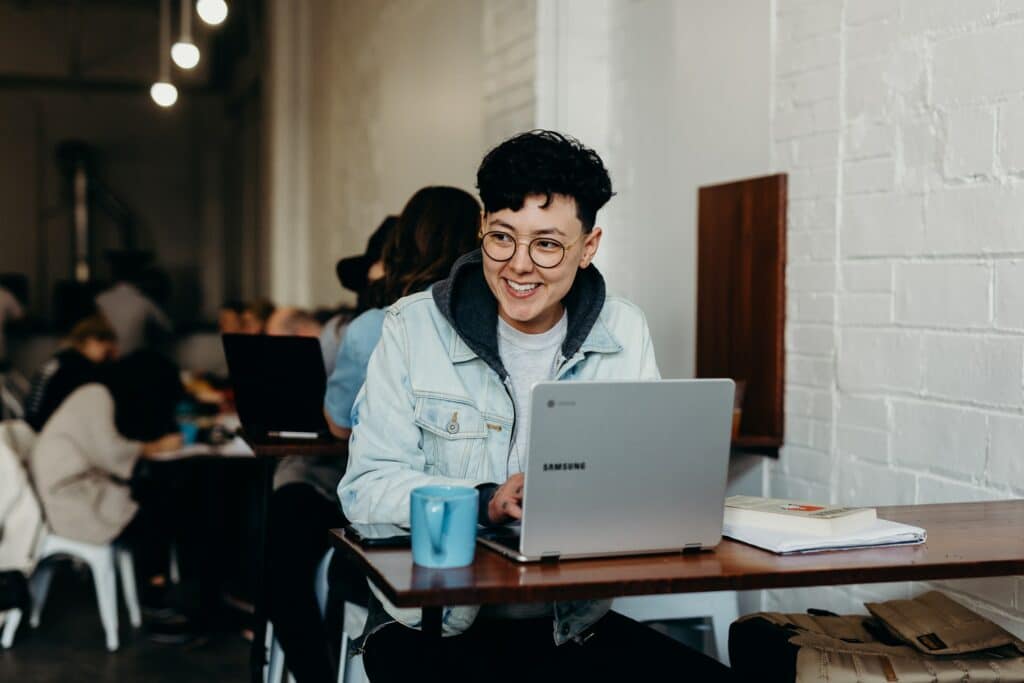 A person sat at a desk in a cafe using a laptop sending a video message for work with a mug next to them and books on the desk.